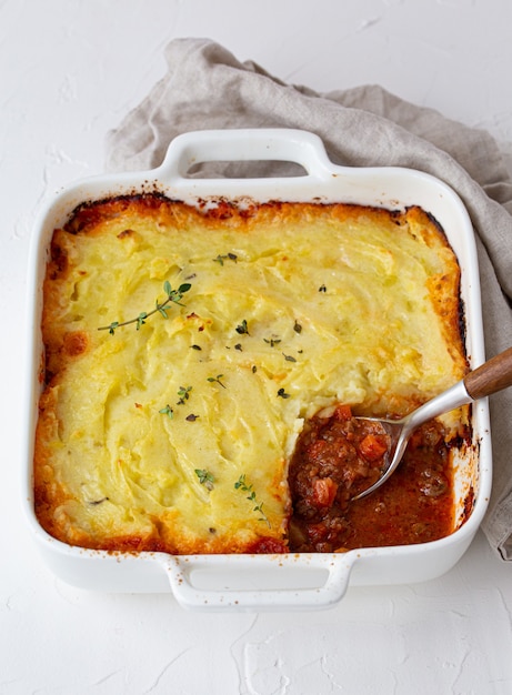 Piatto tradizionale della cucina britannica Casseruola di torta di pastore con carne macinata e purè di patate in teglia in ceramica su tavola rustica bianca con cucchiaio dall'alto