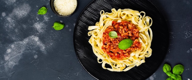 Piatto tradizionale italiano fettuccine con ragù alla bolognese, basilico e parmigiano in banda nera su fondo di legno scuro. Vista dall'alto.