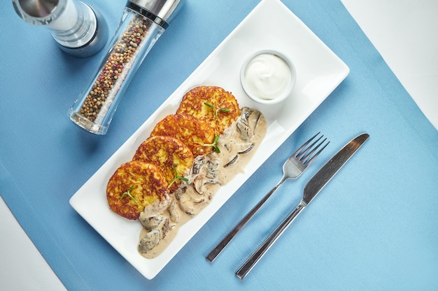 Piatto tradizionale ucraino e bielorusso - frittelle di patate fritte (draniki, deruny) con panna acida e funghi porcini, servite in un piatto bianco su un tavolo blu