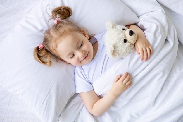 piccola bambina bionda carina che dorme su un letto di cotone bianco a casa abbracciando un orsacchiotto in primo piano