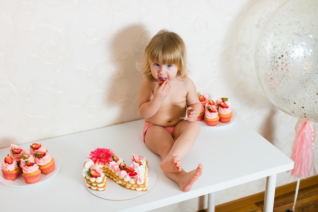 Piccola bambina bionda di due anni in pantaloni rosa che si siede sul tavolo bianco vicino alla sua torta di compleanno e diversi dolci rosa sul tavolo