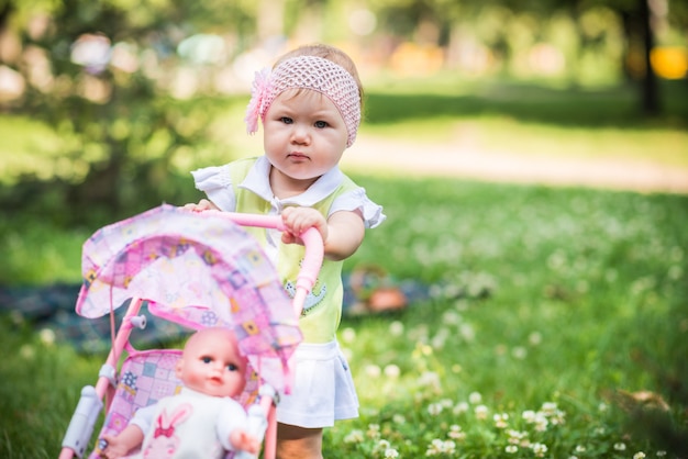 Piccola bambina che cammina sull'erba e passeggino giocattolo a rotazione con la bambola