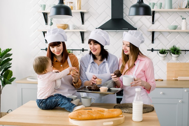 Piccola bambina è seduta sul tavolo in cucina e divertirsi. La nonna e le sue figlie bevono caffè e mangiano muffin. Donne felici in grembiuli bianchi che cuociono insieme. Festa della mamma