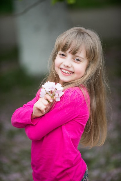 Piccola bambina o bambino carino con adorabile viso sorridente e capelli biondi in camicia rosa che tiene fiore di ciliegio primaverile sakura all'aperto su sfondo sfocato