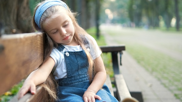 Piccola bambina stanca seduta su una panchina con gli occhi chiusi che riposa nel parco estivo.