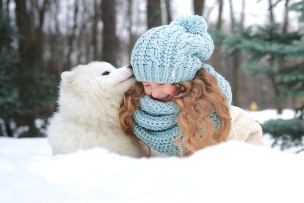 Piccola bella ragazza carina felice bambino positivo bambino è sdraiato sulla neve giocare divertendosi con il suo grande cane bianco al giorno d'inverno nel parco o nella foresta