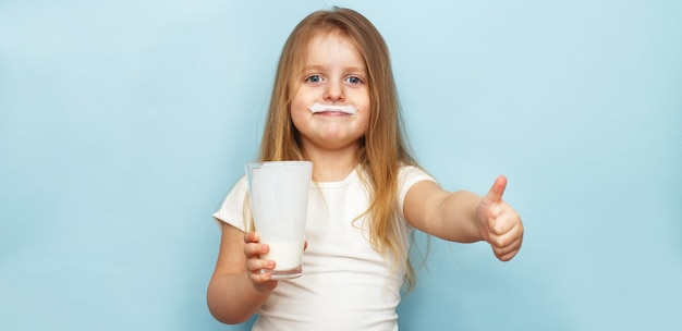 Piccola bella ragazza che tiene il latte in un bicchiere su sfondo blu. bambino beve yogurt. striscione