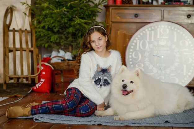 Piccola bella ragazza e un grande cane lanuginoso bianco accanto all'albero di Natale nell'interno del nuovo anno.