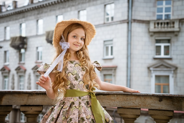 Piccola bella ragazza in vestito e cappello in posa sul balcone della città