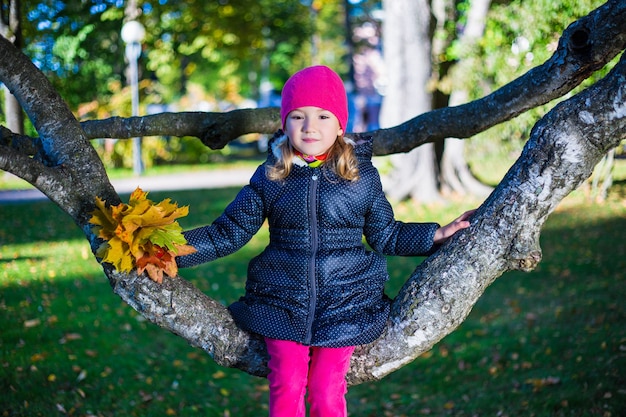 Piccola bella ragazza seduta sul ramo di un albero nel parco autunnale