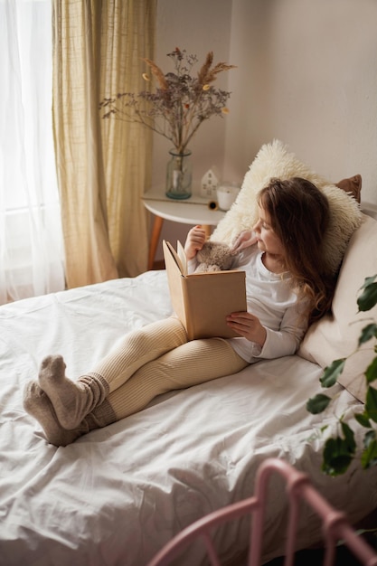 Piccola bellissima ragazza riccia abbraccia il suo coniglio di peluche e legge un libro nel suo letto casa accogliente