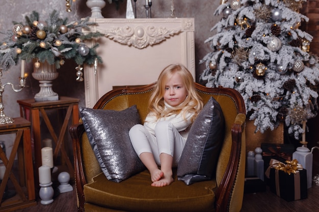 Piccola bionda affascinante si siede avvolta in un tappeto su una bellissima vecchia sedia sotto l'albero di Natale.
