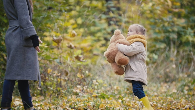 Piccola figlia con sua madre e l'orsacchiotto che camminano nel parco autunnale, teleobiettivo