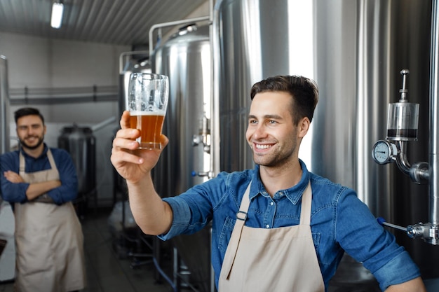 Piccola impresa familiare. Birraio professionista con produzione artigianale di alcolici. Felice millenario orgoglioso bell'uomo in grembiule controlla la qualità della birra vicino a un grande serbatoio di metallo con bevanda artigianale, spazio vuoto