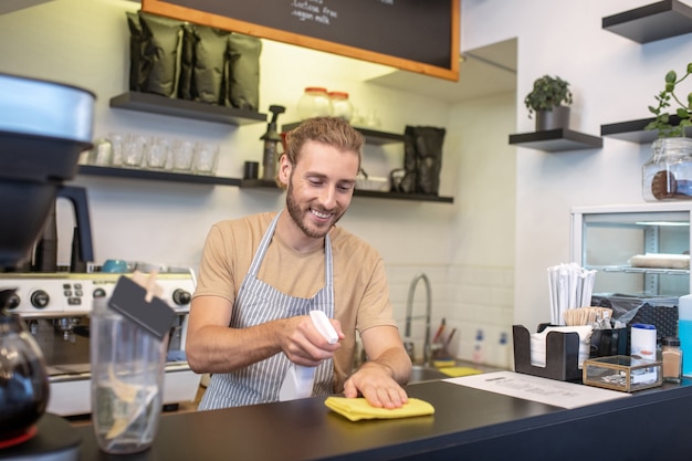 Piccola impresa. Giovane uomo barbuto che si rallegra che pulisce la superficie del bancone del bar nella sua caffetteria