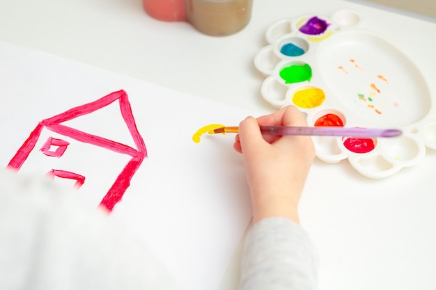 Piccola mano di bambino che disegna la casa e il sole con gli acquerelli. Istruzione e concetto di scuola.