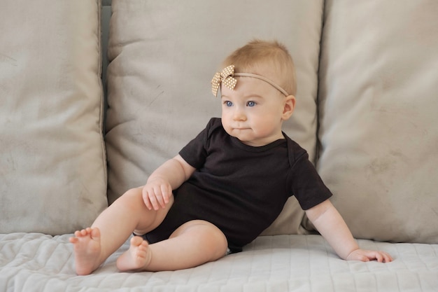 Piccola neonata bionda caucasica seria divertente sveglia, occhi azzurri, seduto sulla superficie dei cuscini del divano morbido beige in corpo nero con fiocco marrone, nastro