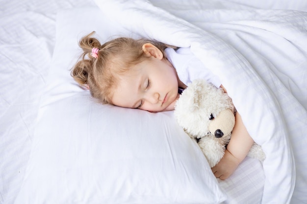 Piccola neonata bionda sveglia che dorme su un letto di cotone bianco a casa che abbraccia un primo piano dell'orsacchiotto