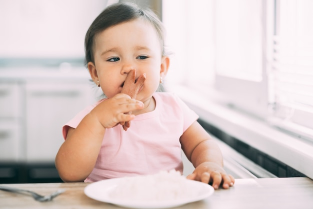 Piccola neonata in cucina che mangia salsiccia e purè di patate