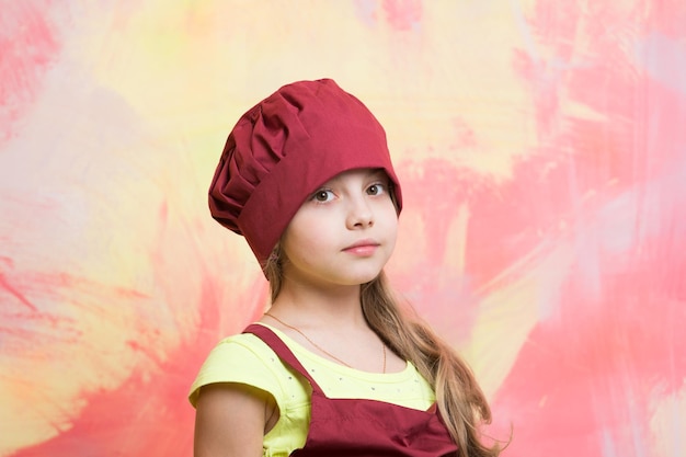 Piccola neonata o bambino carino con viso adorabile in cappello da cuoco rosso e grembiule da cuoco su sfondo astratto colorato