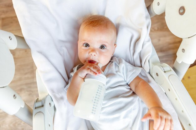 Piccola neonata sveglia che beve latte dalla bottiglia e che guarda l'obbiettivo su sfondo bianco neonato ...