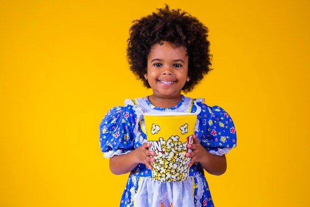 Piccola ragazza afro carina in abito festa junina che mangia popcorn su sfondo giallo