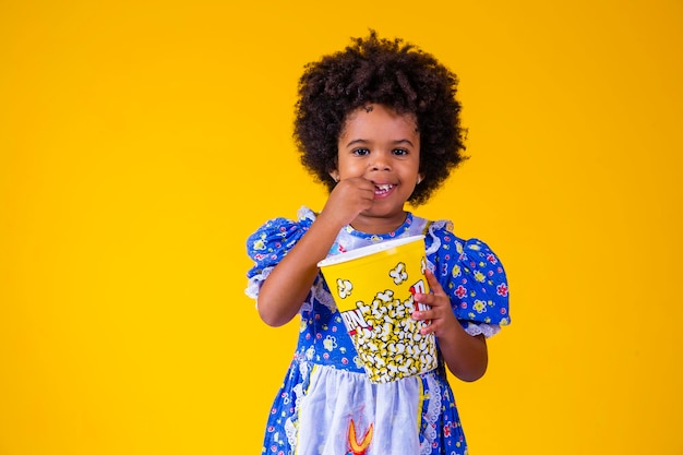 Piccola ragazza afro carina in abito festa junina che mangia popcorn su sfondo giallo