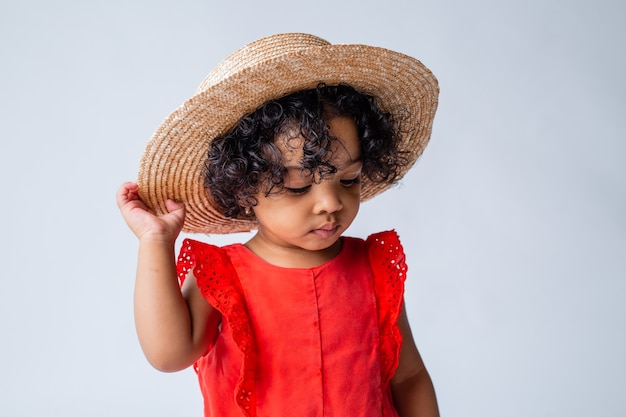 piccola ragazza afroamericana in abiti estivi rossi e un cappello di paglia su uno sfondo bianco in studio