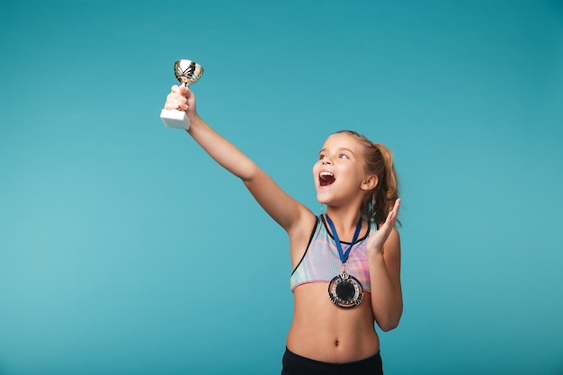 Piccola ragazza allegra di sport che celebra la vittoria isolata sopra la parete blu, che porta una medaglia d'oro, che mostra un trofeo