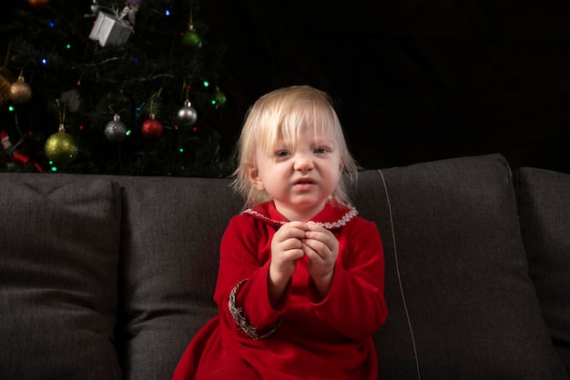 Piccola ragazza arruffata in un vestito rosso si siede sul divano sullo sfondo di un albero di Natale Natale e capodanno