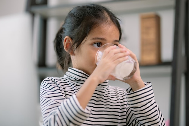 Piccola ragazza asiatica carina che beve latte al tavolo a casa si diverte a bere latte