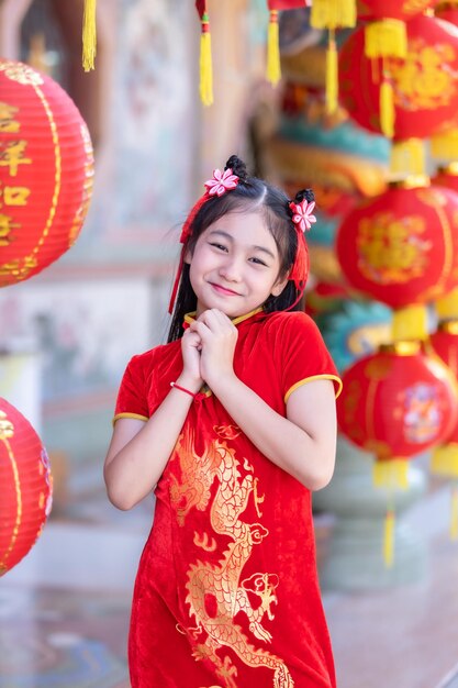 Piccola ragazza asiatica carina che indossa il cheongsam cinese tradizionale rosso con lanterne di carta con l'alfabeto cinese benedizioni scritte su di esso è una decorazione di complimento per la benedizione della fortuna per il capodanno cinese