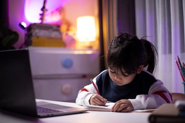 Piccola ragazza asiatica che fa i compiti con il computer portatile