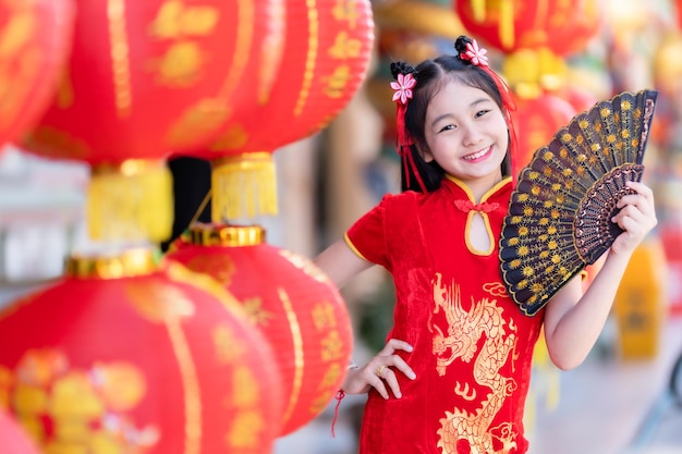 Piccola ragazza asiatica che indossa un cheongsam cinese tradizionale rosso e tiene in mano una lanterna Fanningand con l'alfabeto cinese Le benedizioni scritte su di esso sono una decorazione di complimento per la benedizione della fortuna per il nuovo anno