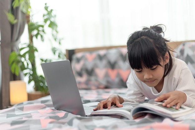 Piccola ragazza asiatica che usa un computer portatile mentre è sdraiato sul letto