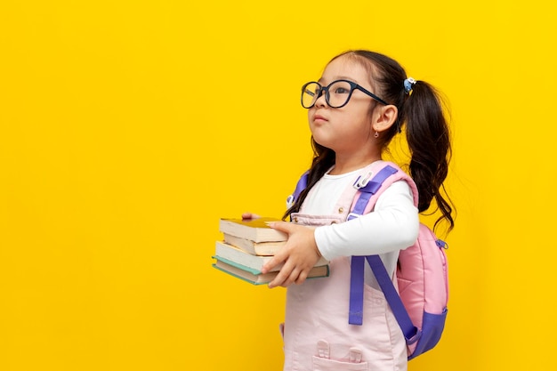 Piccola ragazza asiatica con gli occhiali e con zaino che tiene libri su sfondo giallo