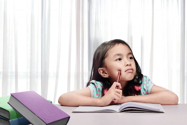 Piccola ragazza asiatica con libro e penna pensando quando si fa i compiti