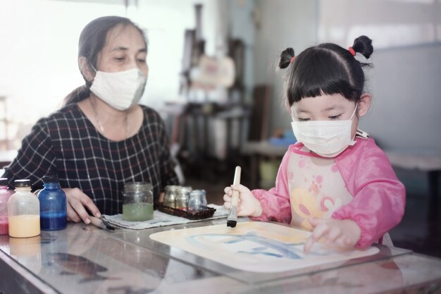 Piccola ragazza asiatica con maschera igienica facciale e madre che dipinge colori ad acqua insieme a casa, messa a fuoco selettiva. Quarantena, isolamento domiciliare durante la pandemia di COVID-19.