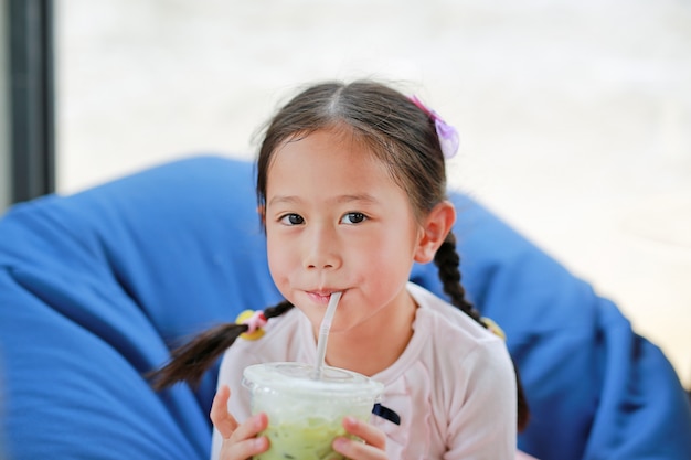 Piccola ragazza asiatica felice del bambino che beve il tè verde ghiacciato di Matcha che si trova sul sofà al caffè.