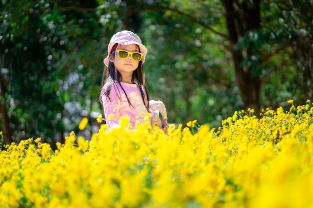 Piccola ragazza asiatica in cappello rosa ed occhiali da sole di usura del vestito che stanno nel giardino floreale giallo