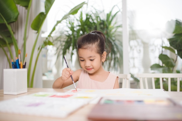 Piccola ragazza asiatica sveglia che gode della pittura a casa con l'acquerello di carta e la spazzola di arte