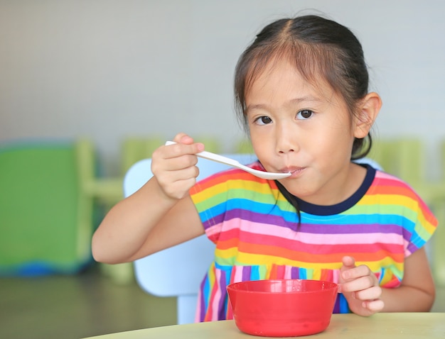 Piccola ragazza asiatica sveglia che mangia cereale con i fiocchi di granturco e latte sulla tavola