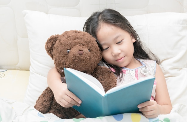 Piccola ragazza asiatica sveglia in camicia da notte che legge un libro con orsacchiotto sul letto