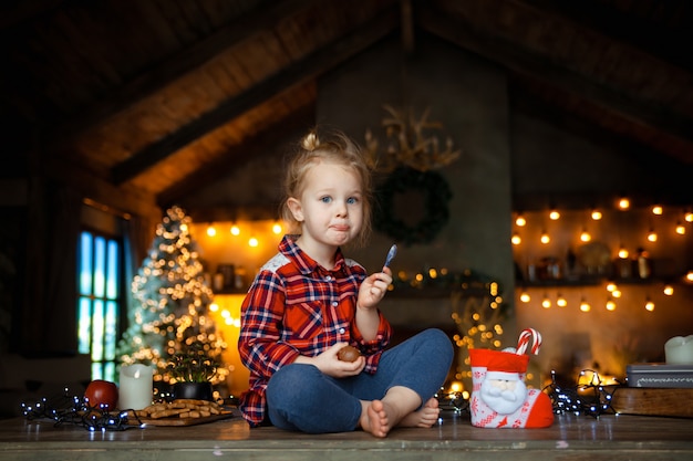 Piccola ragazza bionda bianca che si siede sulla tavola di legno e che mangia un uovo di cioccolato dal suo regalo di natale.