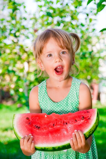 Piccola ragazza bionda con l'anguria della grande fetta nell'ora legale in parco, all'aperto. Sorpresa bambino Oops, wow.