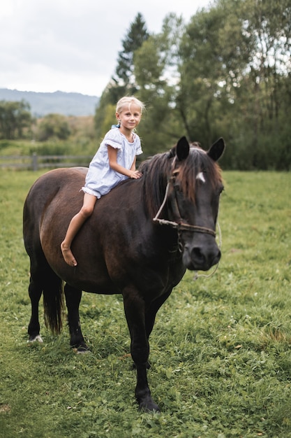 Piccola ragazza bionda del bambino in vestito che monta un cavallo.