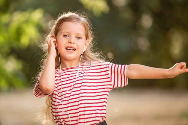 Piccola ragazza bionda felice che ascolta la musica con le cuffie, ballando e cantando in natura nel parco.