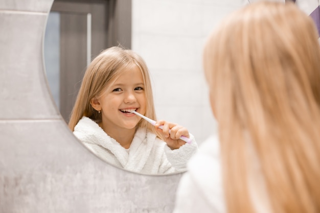 piccola ragazza bionda in un accappatoio bianco si lava i denti davanti allo specchio del bagno