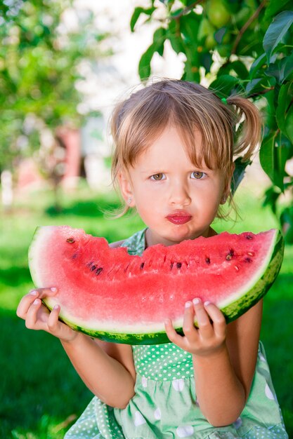 Piccola ragazza bionda seria con l'anguria della grande fetta nell'ora legale in parco, all'aperto.