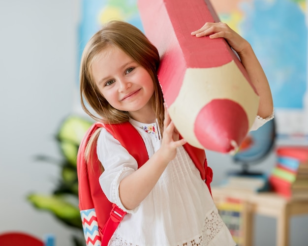 Piccola ragazza bionda sorridente con la borsa di rd che tiene matita decorativa rossa enorme nella classe di scuola